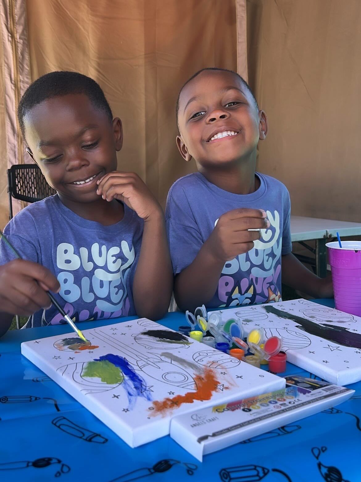 Two children painting at a table, smiling beside a tray of colorful art supplies.