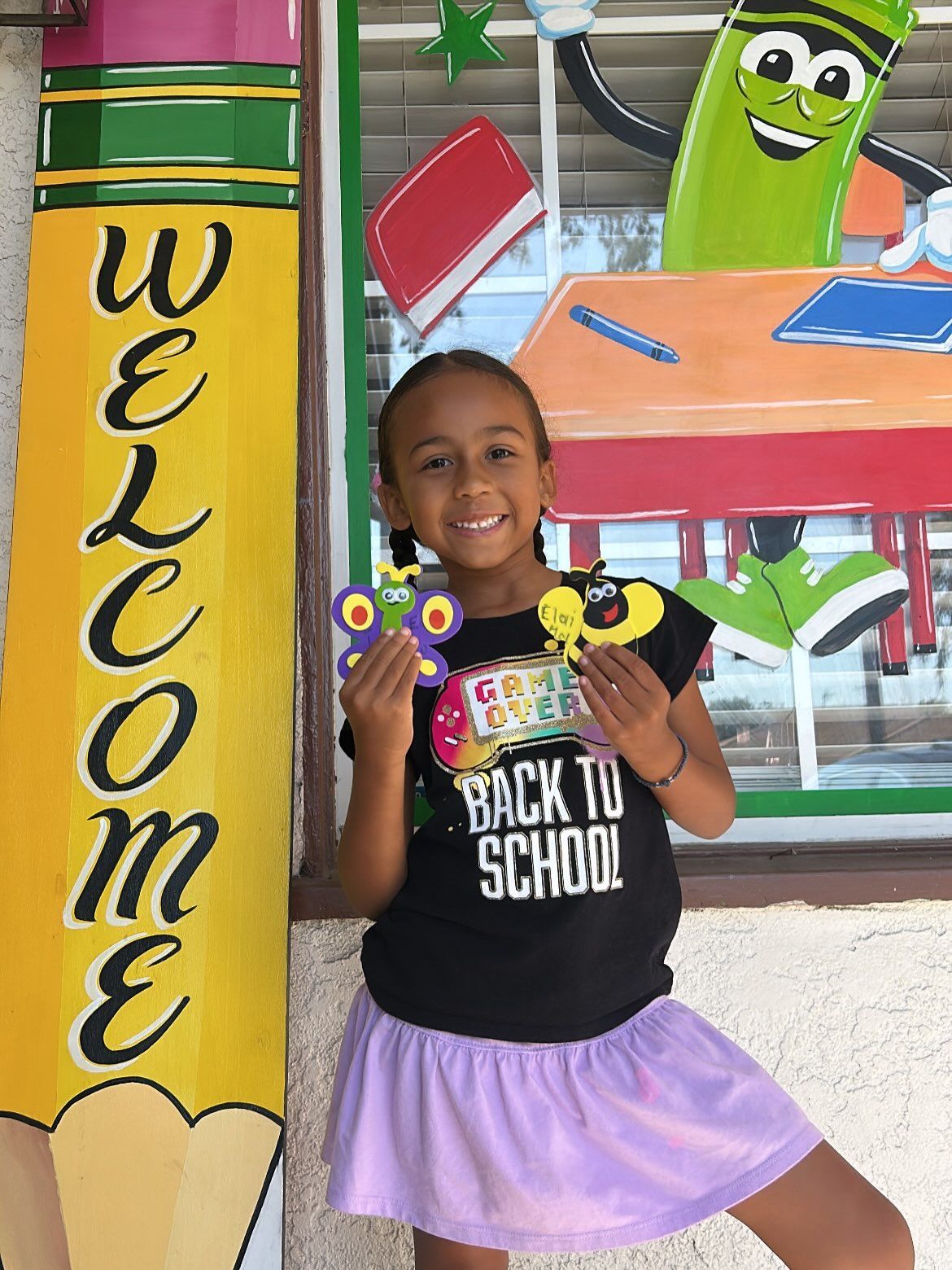 Girl posing by a yellow “Welcome” pillar, holding decorations in front of a colorful back-to-school display.