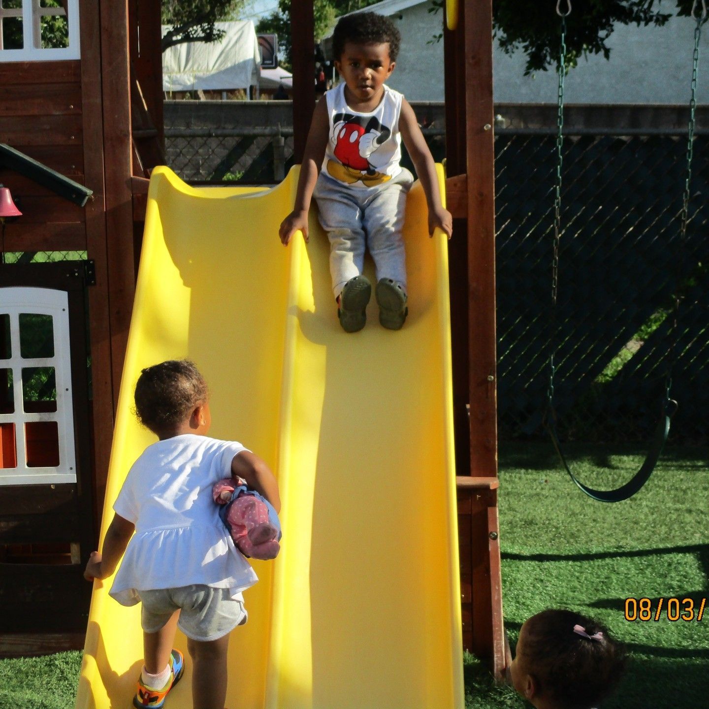 Two children on a yellow playground slide, one climbing up while another sits at the top.