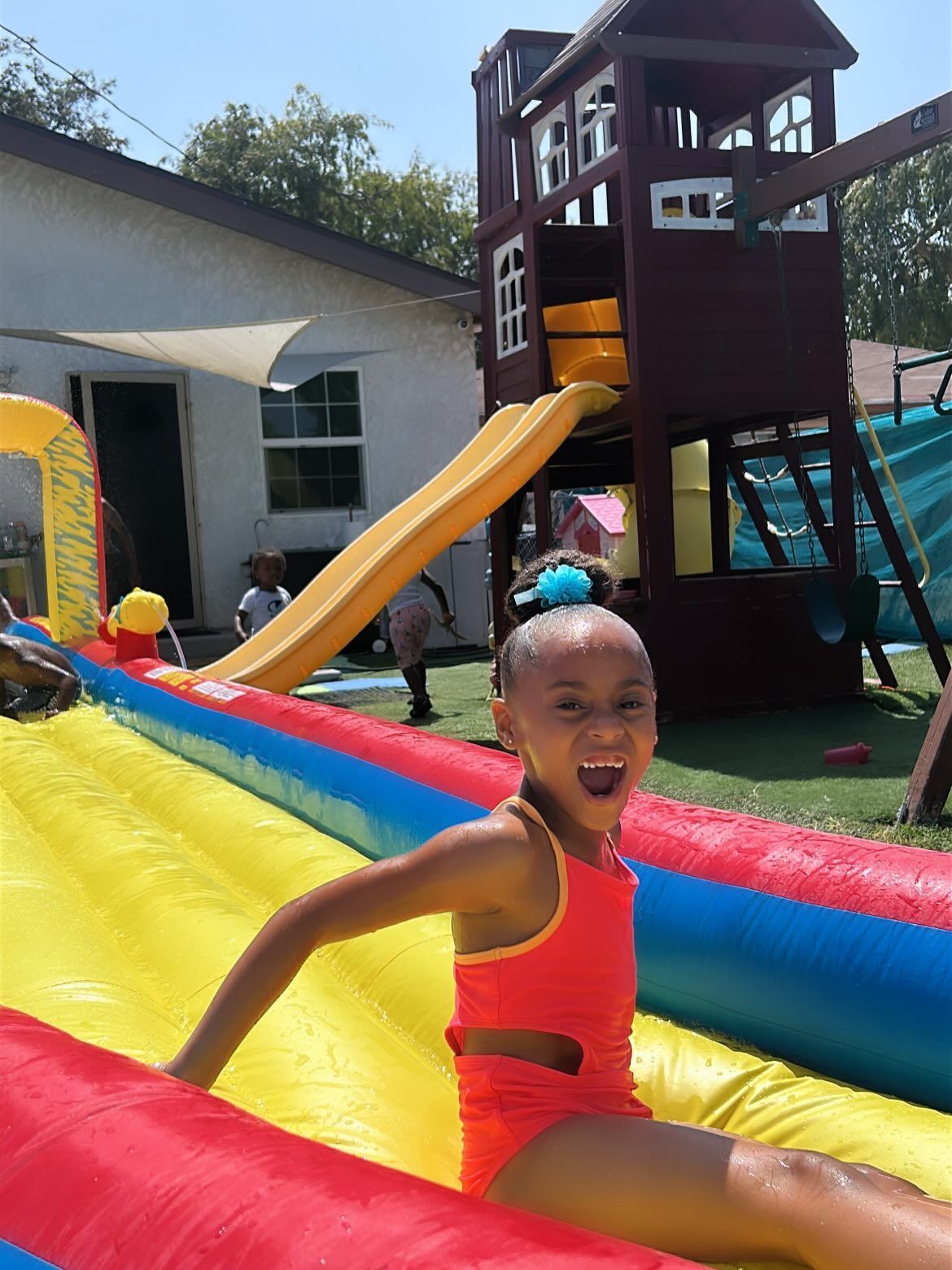 Child in a red swimsuit sliding down a yellow inflatable slide, smiling outdoors near a playground structure