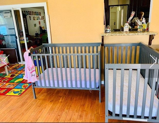 Two gray cribs in a colorful playroom with wood flooring and a kitchen pass-through window