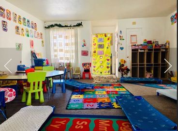 Colorful daycare classroom with rugs, tables, chairs, shelves, and a window with curtains