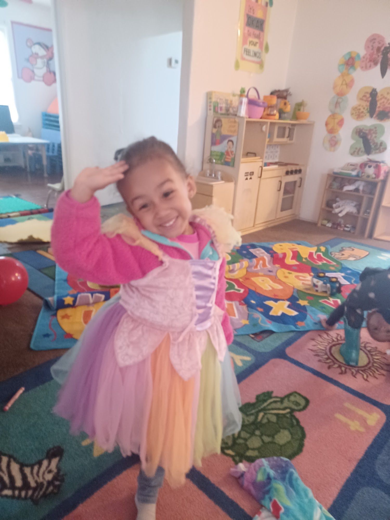 Child in a pink princess dress smiling and posing in a colorful playroom.