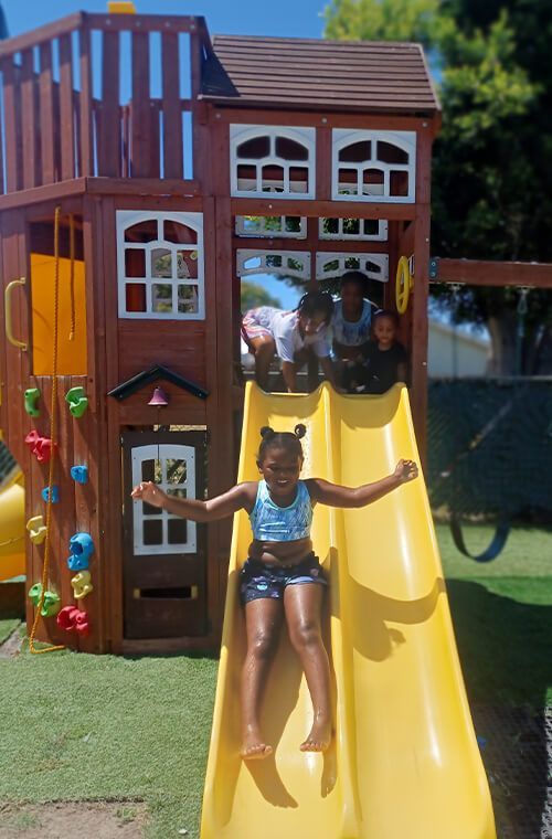 Child sliding down yellow slide from a wooden playhouse in a sunny backyard.