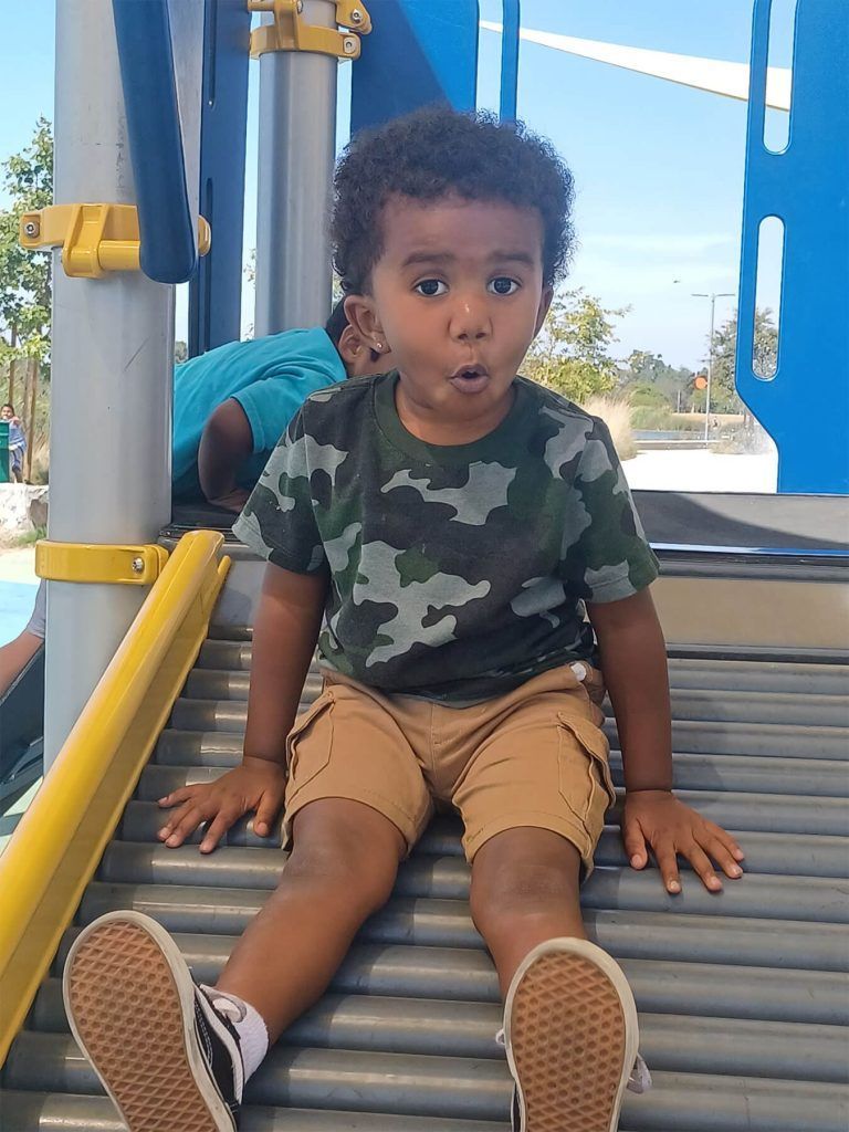 Young child in camouflage shirt and tan shorts sitting on a playground slide, looking surprised