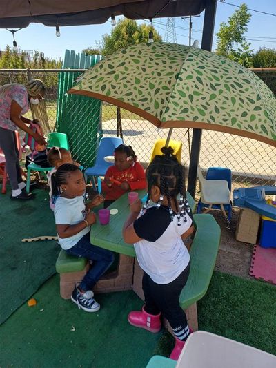 Children sit at a green picnic table under a leaf-patterned umbrella in a sunny outdoor play area