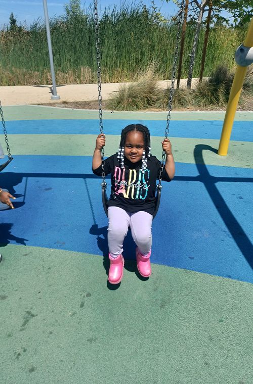 Child smiling on a swing at a colorful playground, wearing a black shirt, pink pants, and pink shoes.