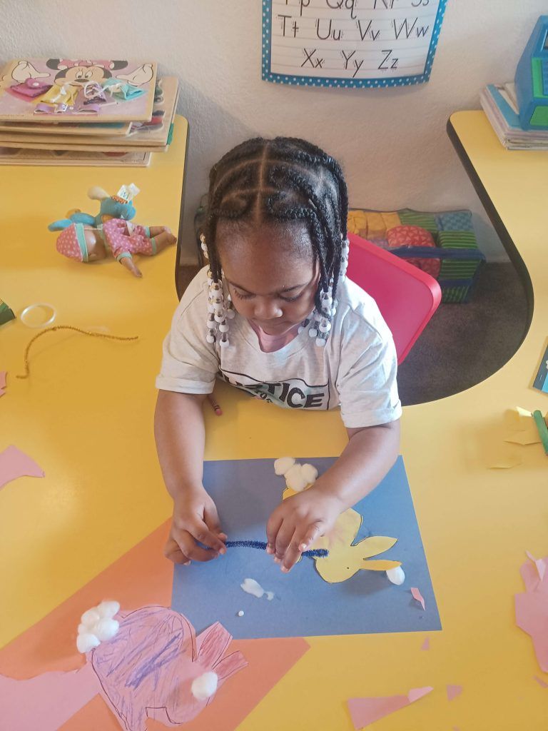 Child at a yellow table making a flower craft on blue paper in a classroom.
