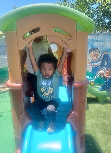 Child on a blue slide in a playground playhouse, holding overhead bars under a green roof.