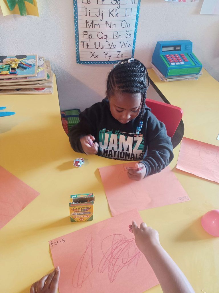 Child coloring at a yellow classroom table with crayons and paper, colorful supplies nearby.