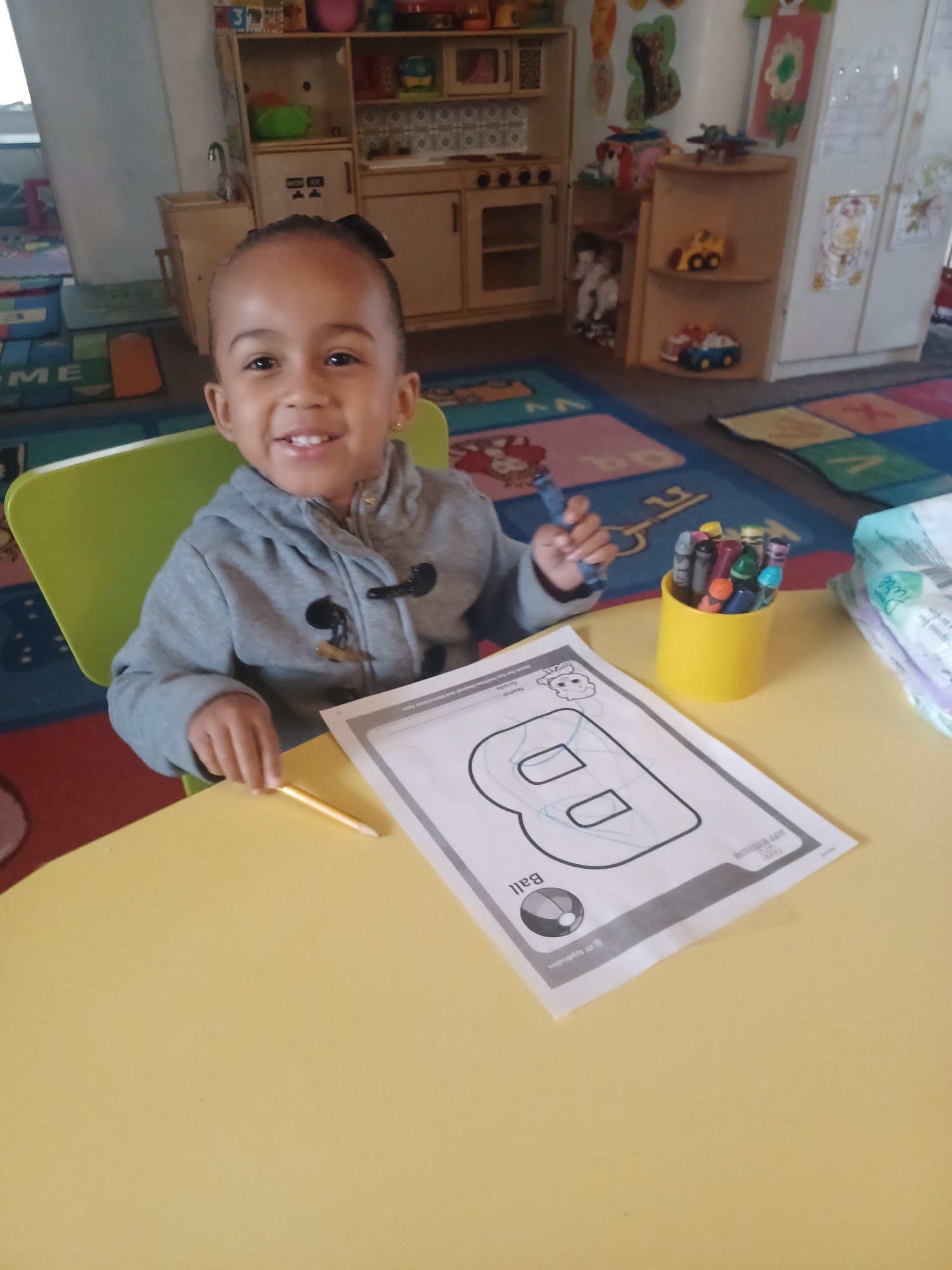 Child smiling at a classroom table with a worksheet and colorful crayons in a classroom.