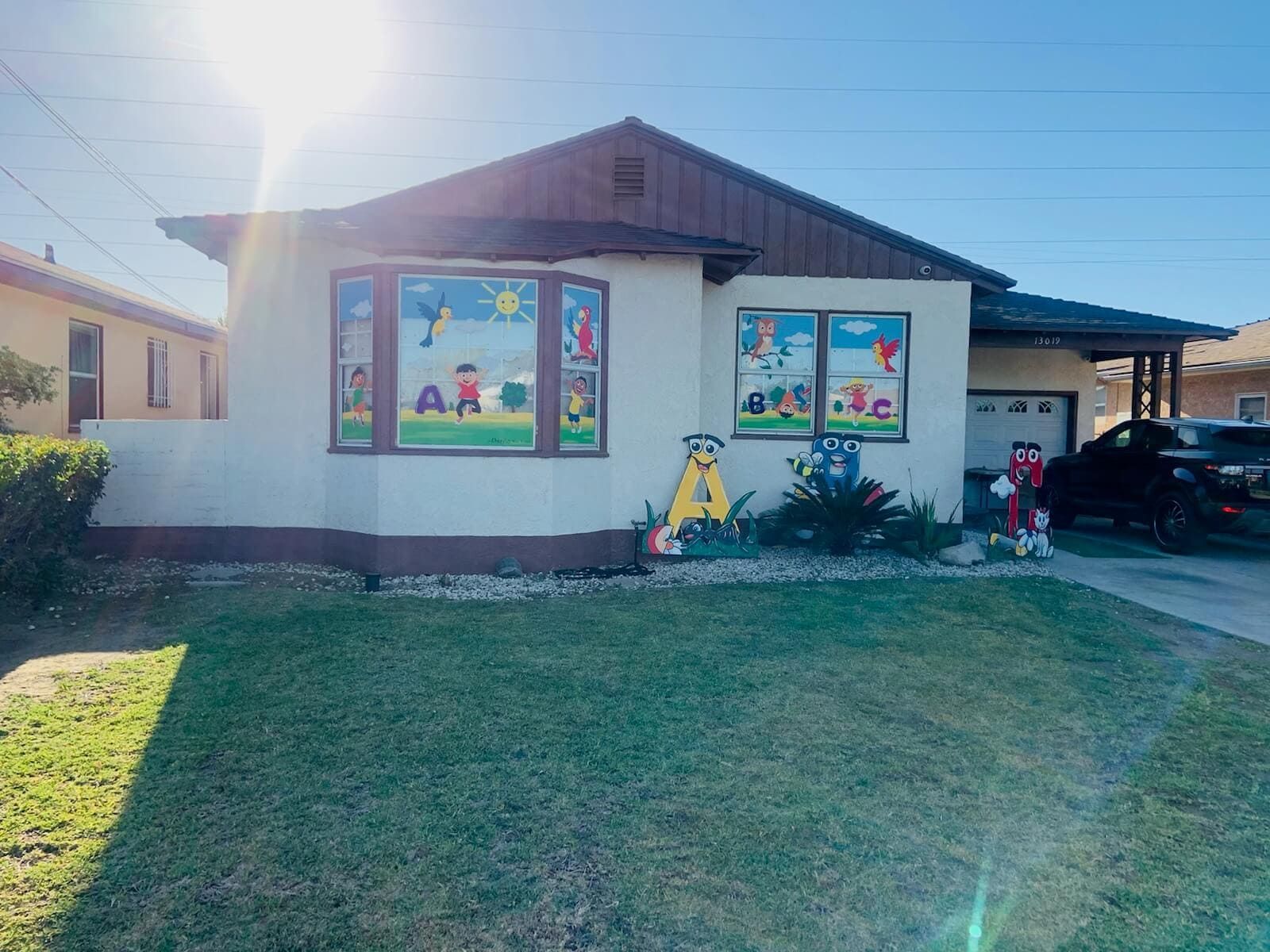 Single-story house with colorful child-themed window art and lawn in front, sun glare at left.