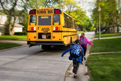 Two children with backpacks walking to a yellow school bus on a suburban street