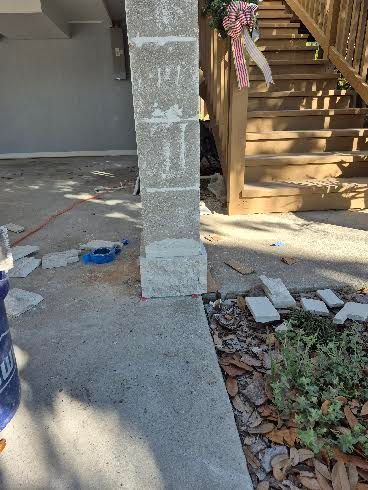 Outdoor walkway beside a porch staircase, with a concrete column, scattered debris, and a blue bucket.