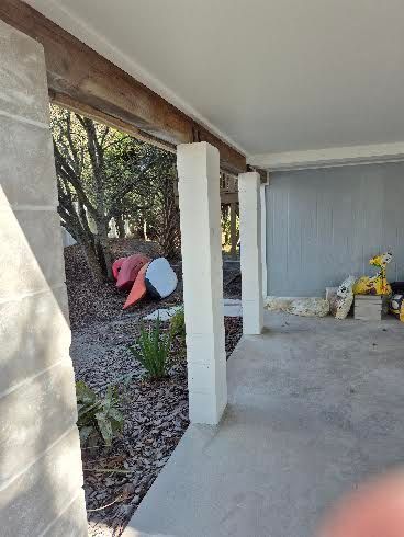 Covered patio with concrete floor, white columns, and a gray wall beside a wooded yard.