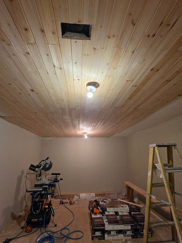 Room under renovation with wood-paneled ceiling, ladder, tools, and construction materials.