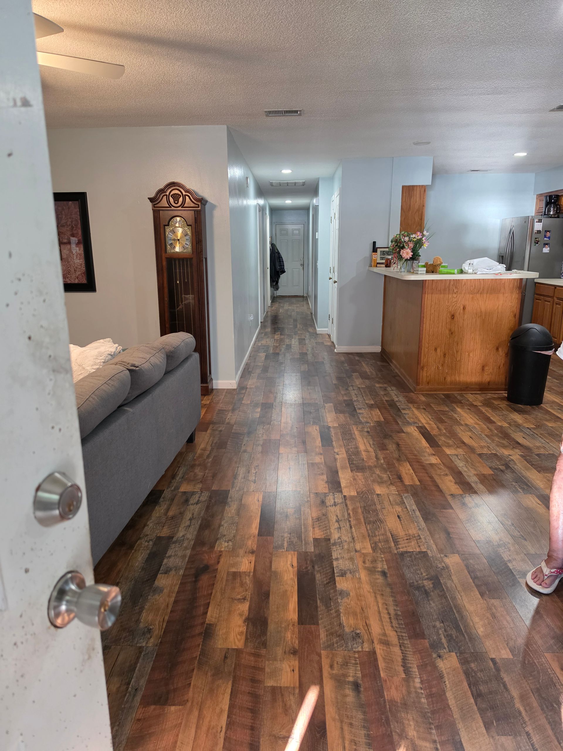 Living room with wood floors, gray sofa, grandfather clock, and open hallway leading back