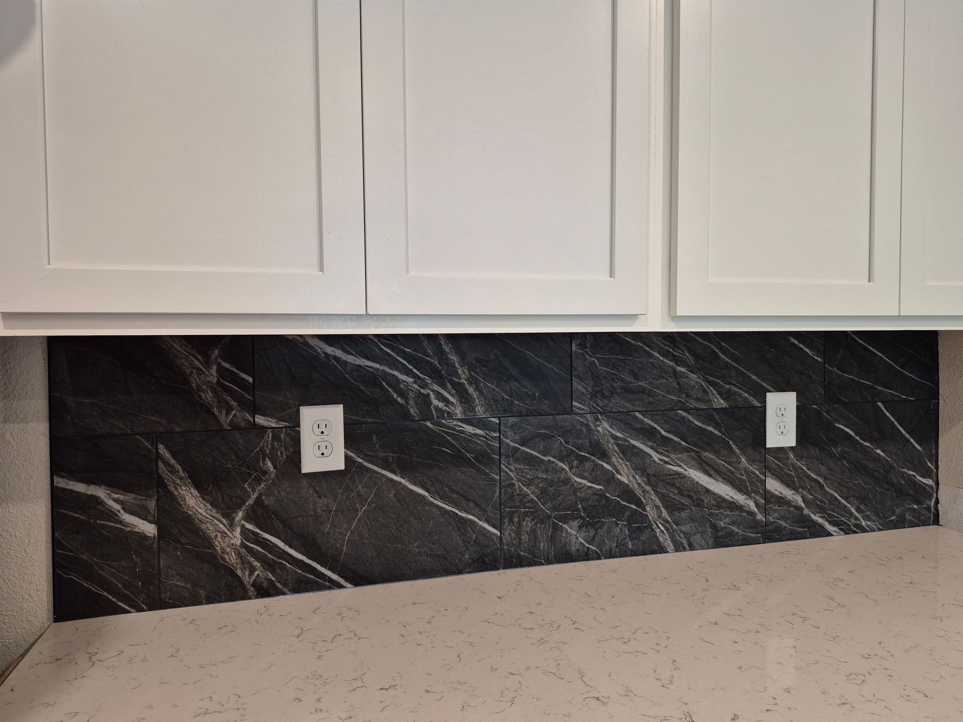 Kitchen backsplash with dark marble-pattern tile, white cabinets, and countertop outlets