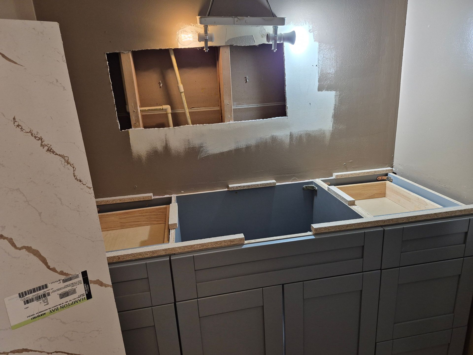 Kitchen under renovation with gray cabinets, unfinished wall cutout, and a marble-look tile panel on the left