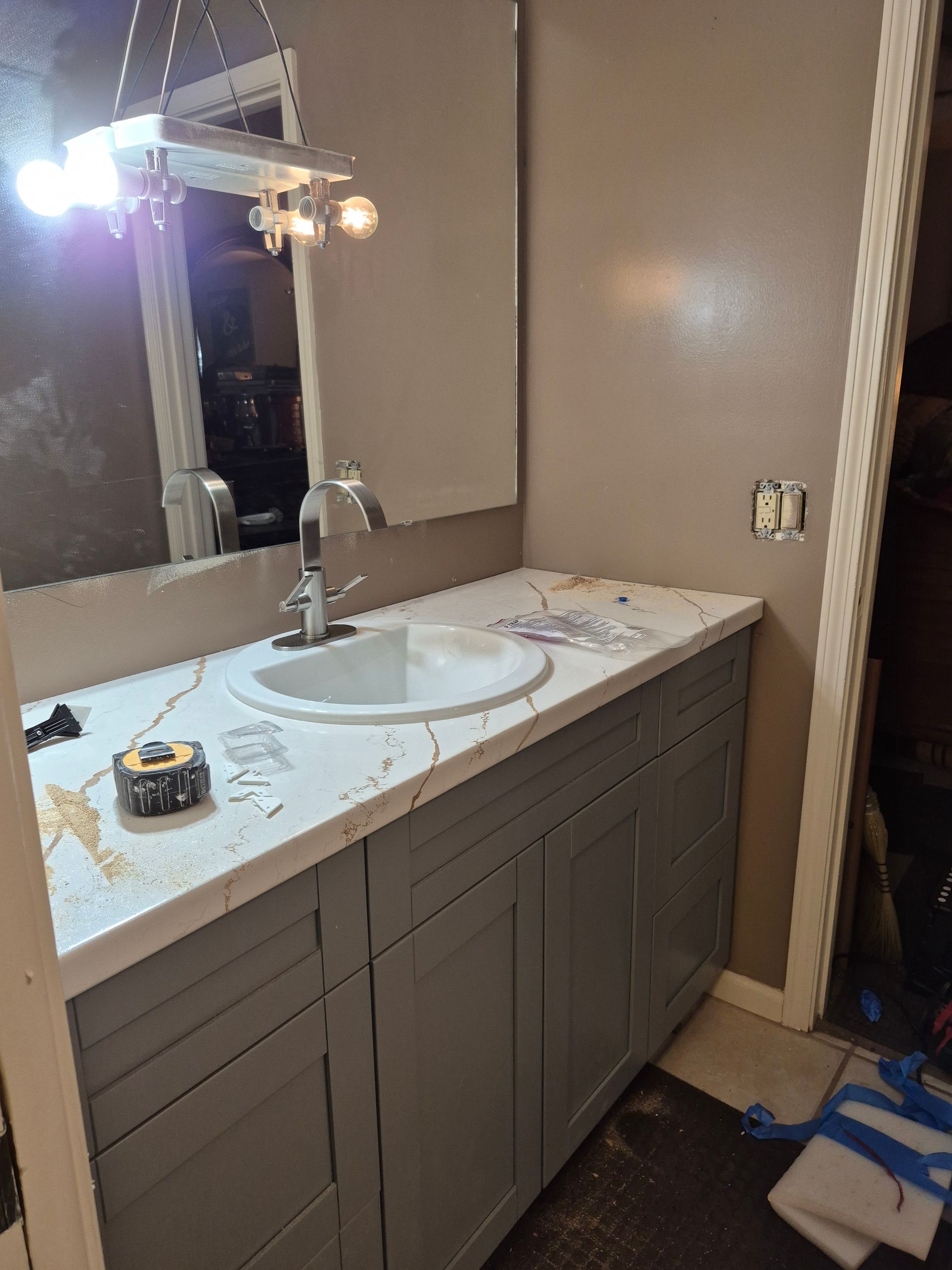 Bathroom vanity with white countertop, large mirror, and gray cabinets; cleaning supplies on the floor.