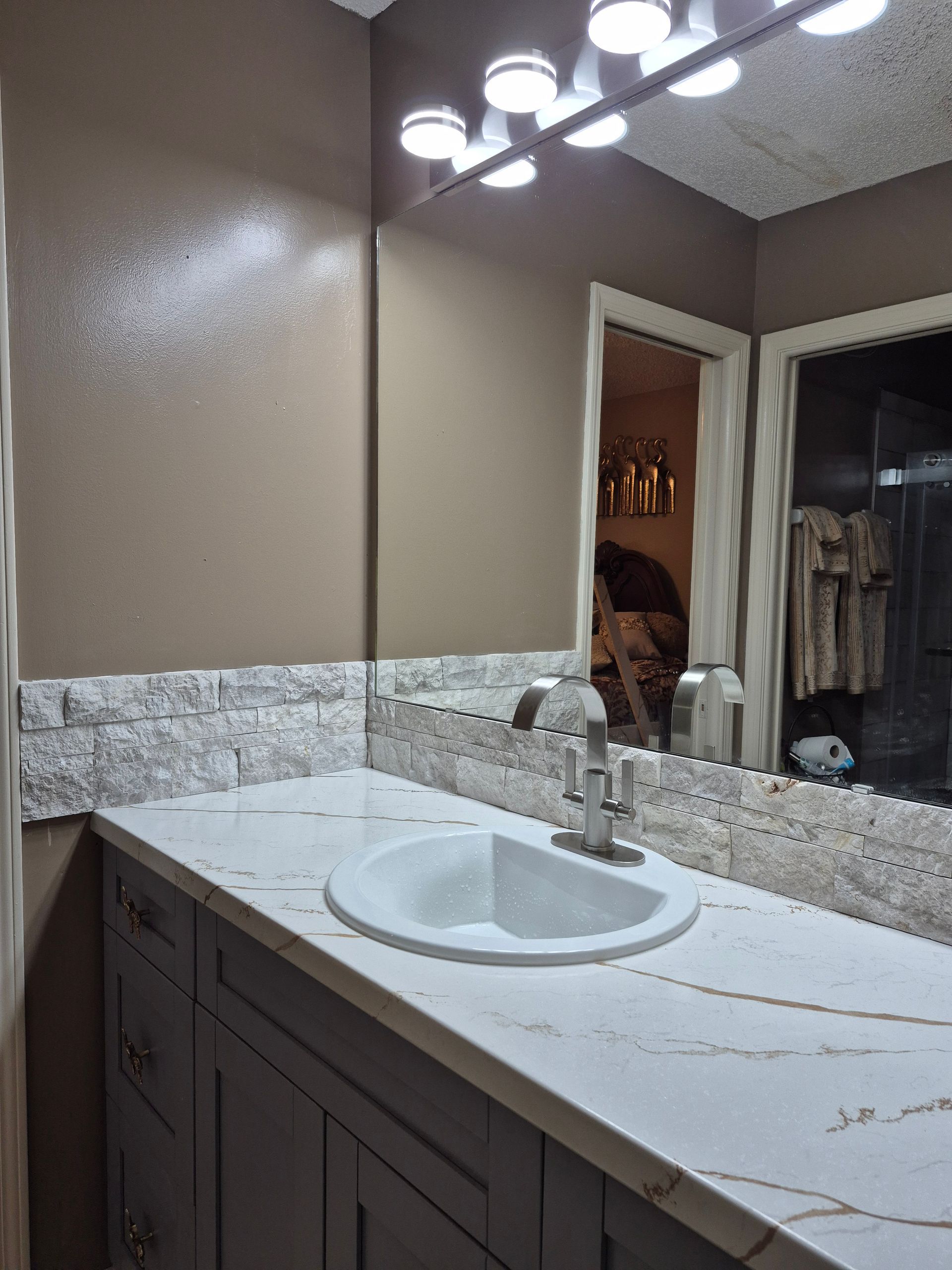 Bathroom vanity with white countertop, round sink, mirror, and overhead lights.