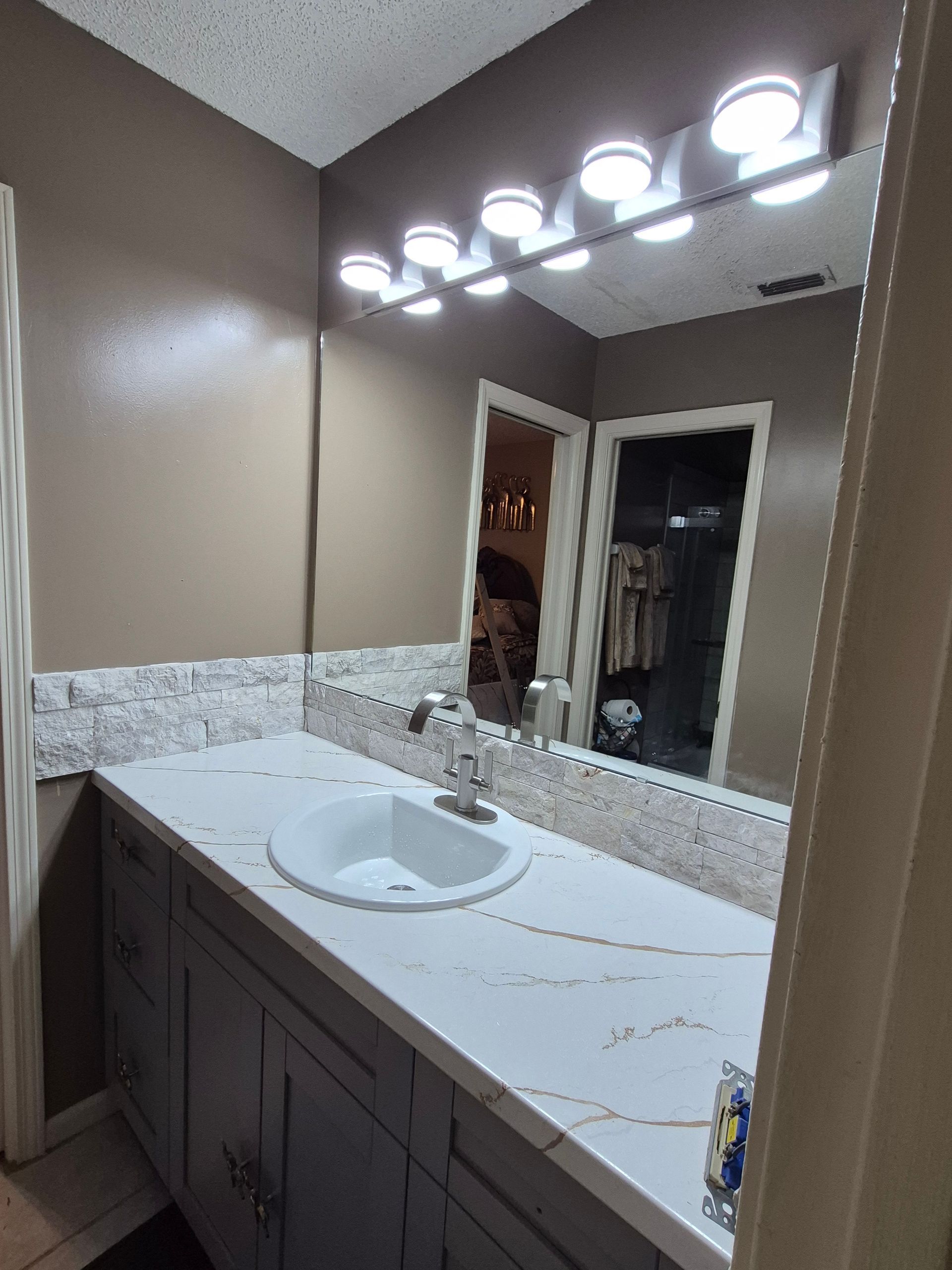 Bathroom vanity with white sink, marble countertop, large mirror, and bright light fixture.