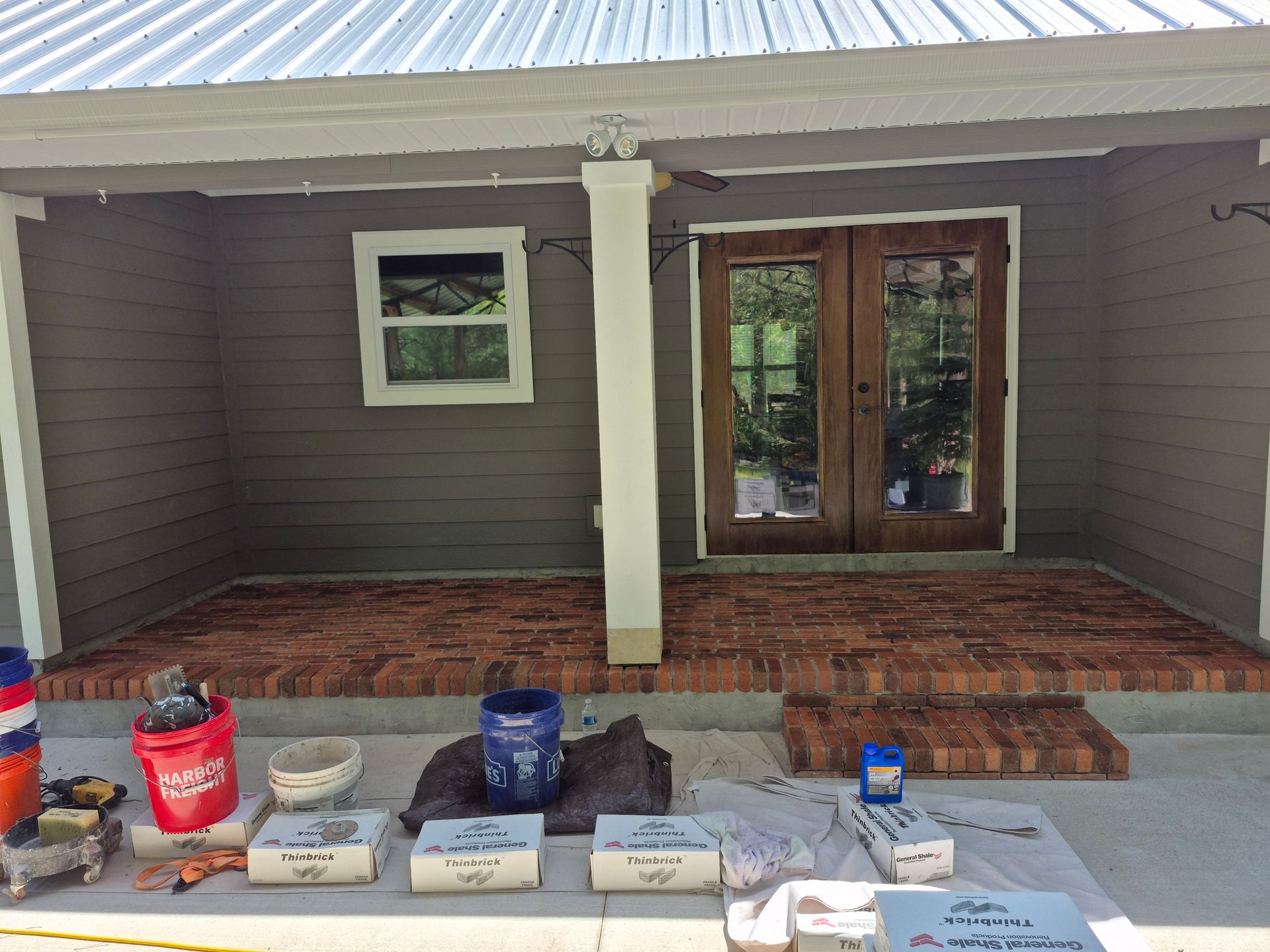 Front porch with tools and buckets set out before double glass doors and a brick step.