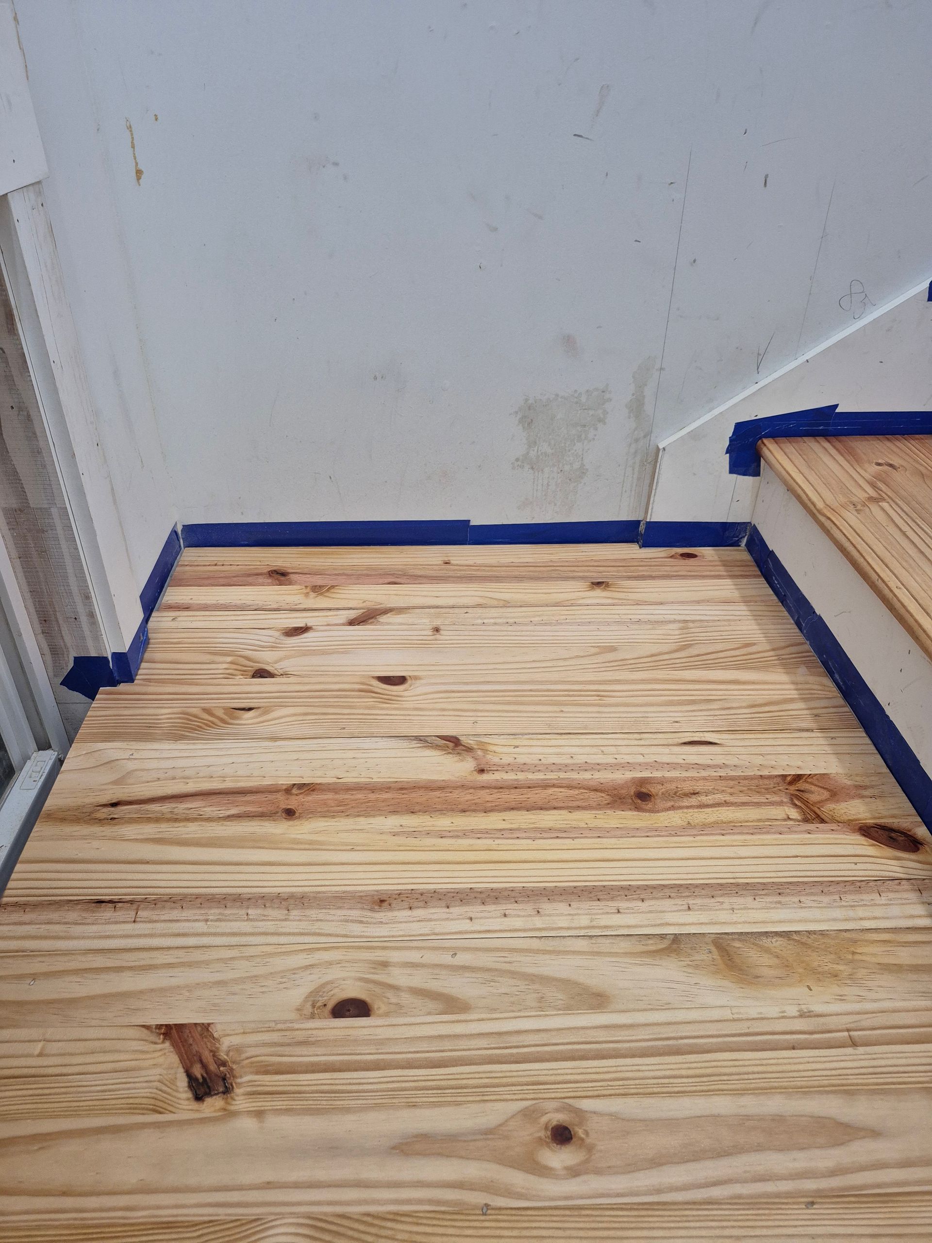 Small unfinished plywood floor in a corner room with blue painter’s tape along the edges.