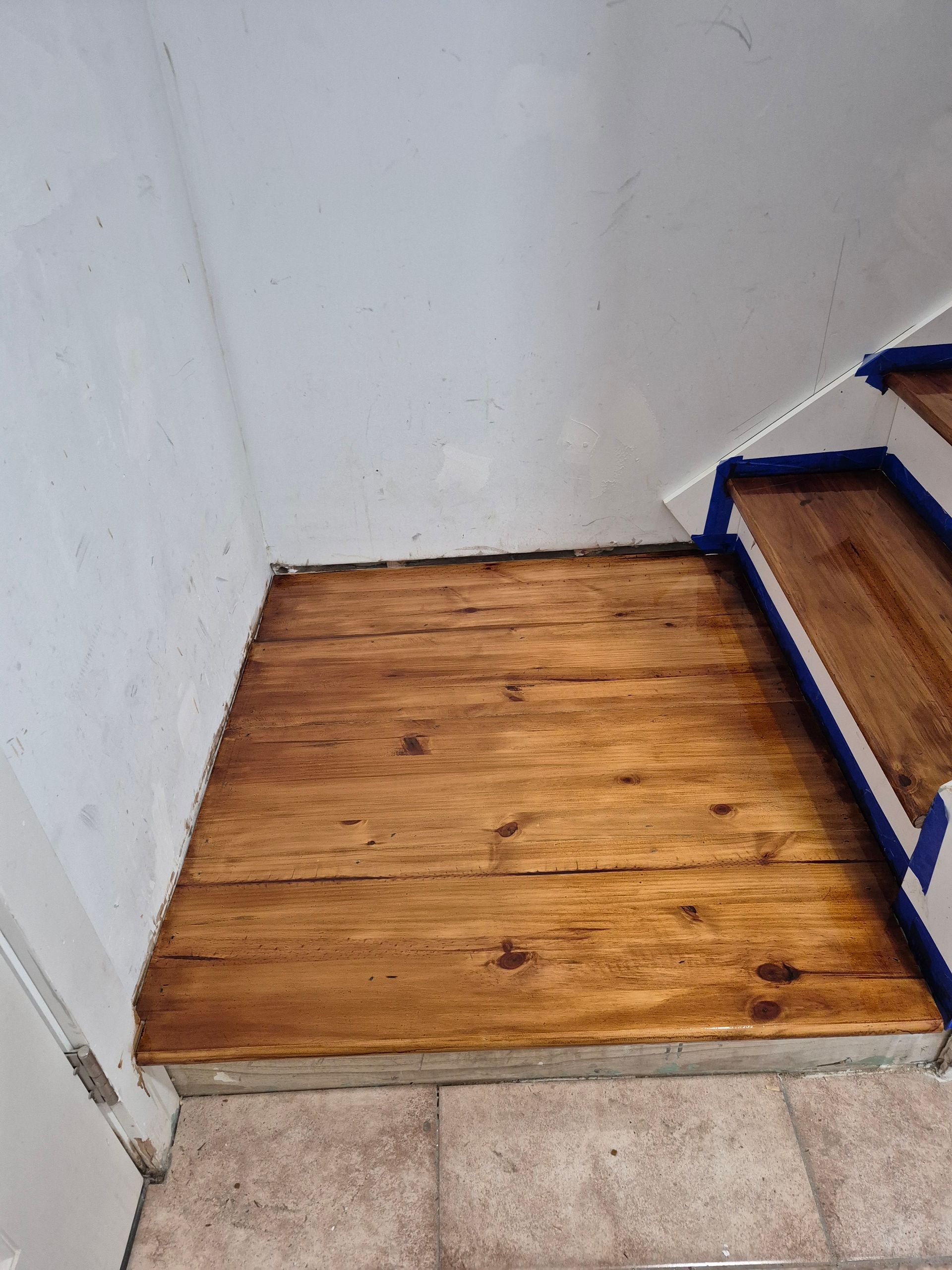 Small wooden platform at the base of a stairway beside a white wall and tiled floor.