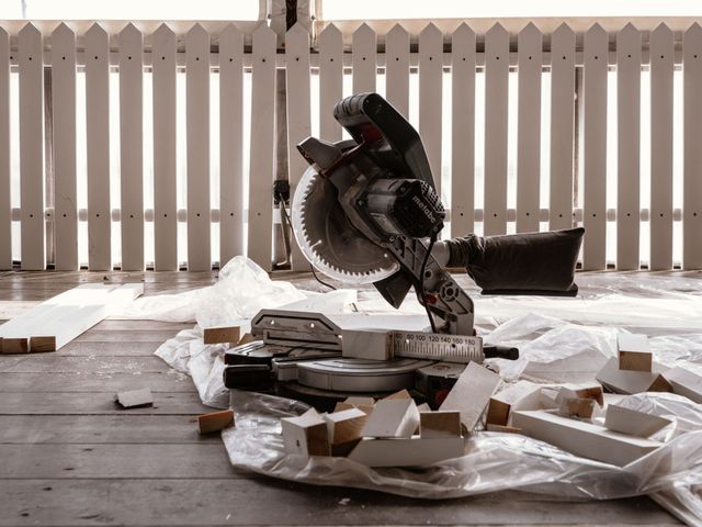 Broken miter saw surrounded by debris in a wood-floored room with a white picket fence backdrop