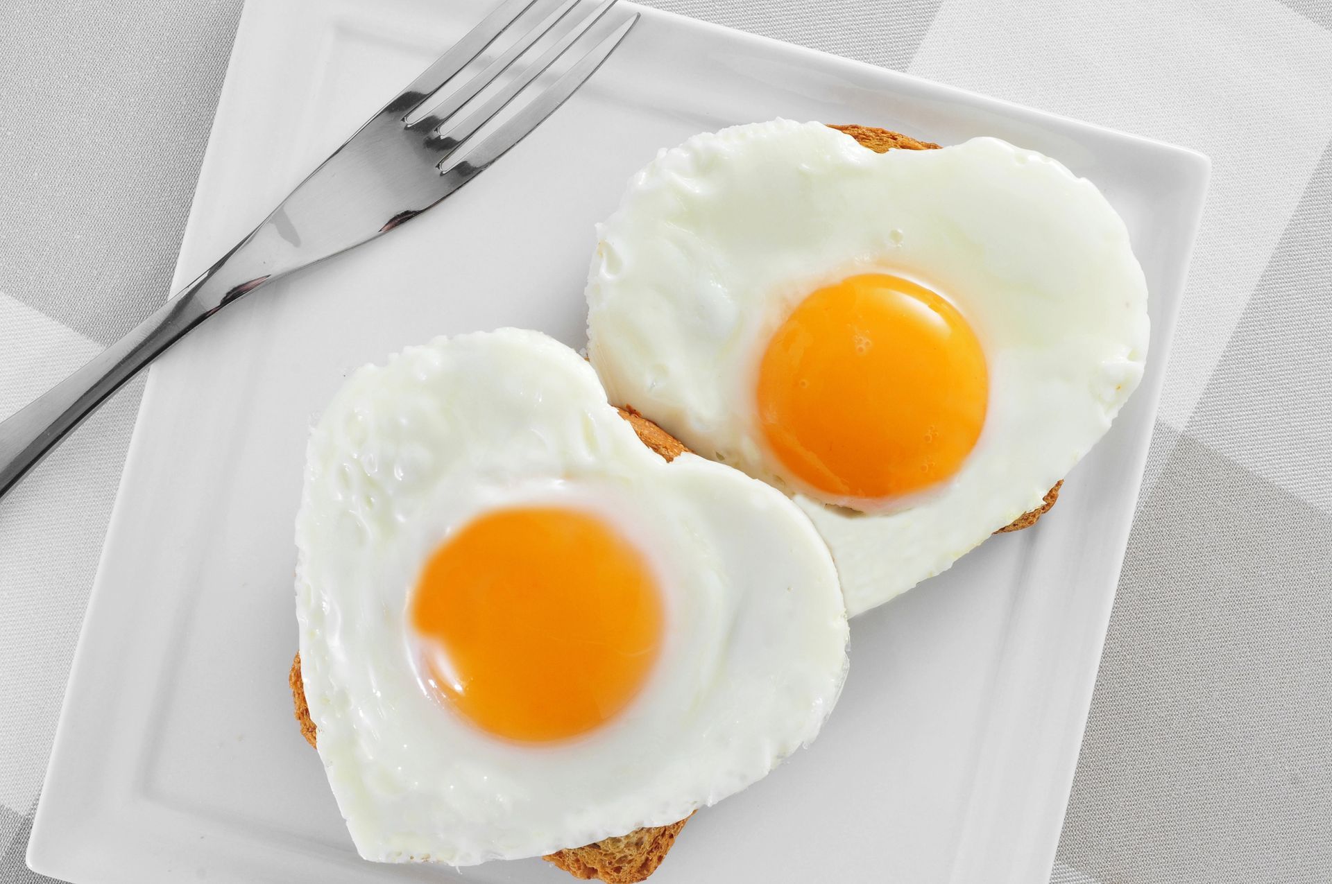 Due uova fritte a forma di cuore su pane tostato, servite su un piatto bianco con una forchetta, sopra una superficie a scacchi.