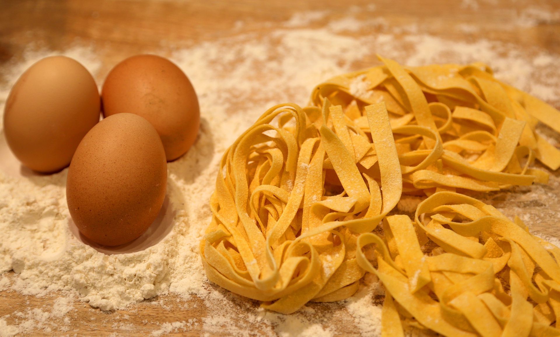 Tre uova marroni accanto a una pila di tagliatelle gialle, su una superficie di legno infarinata.