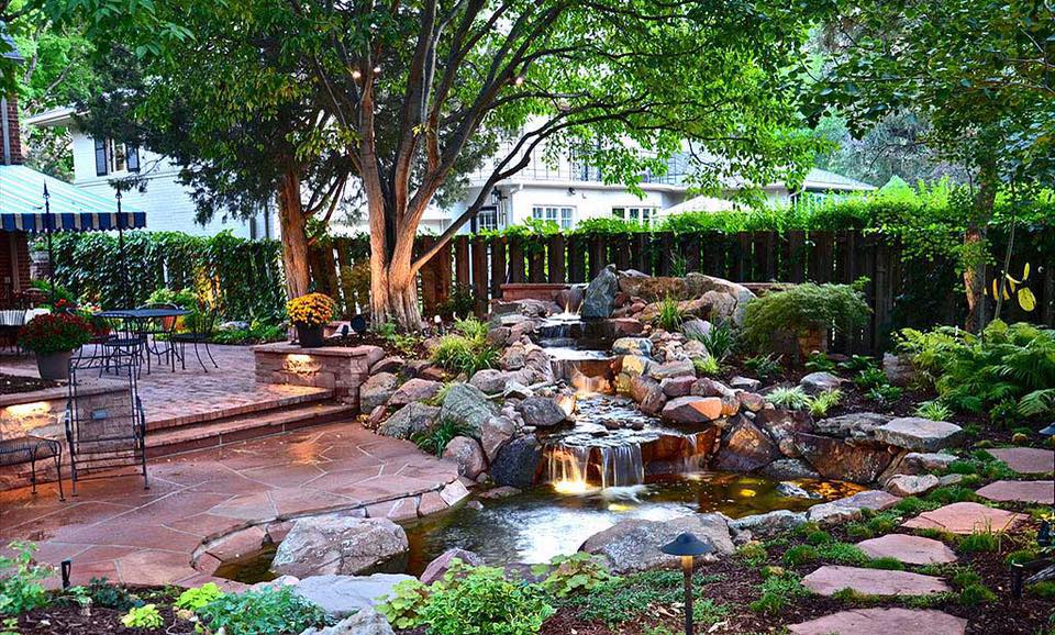 A lush backyard with a stone patio, waterfall, pond, and surrounding greenery.