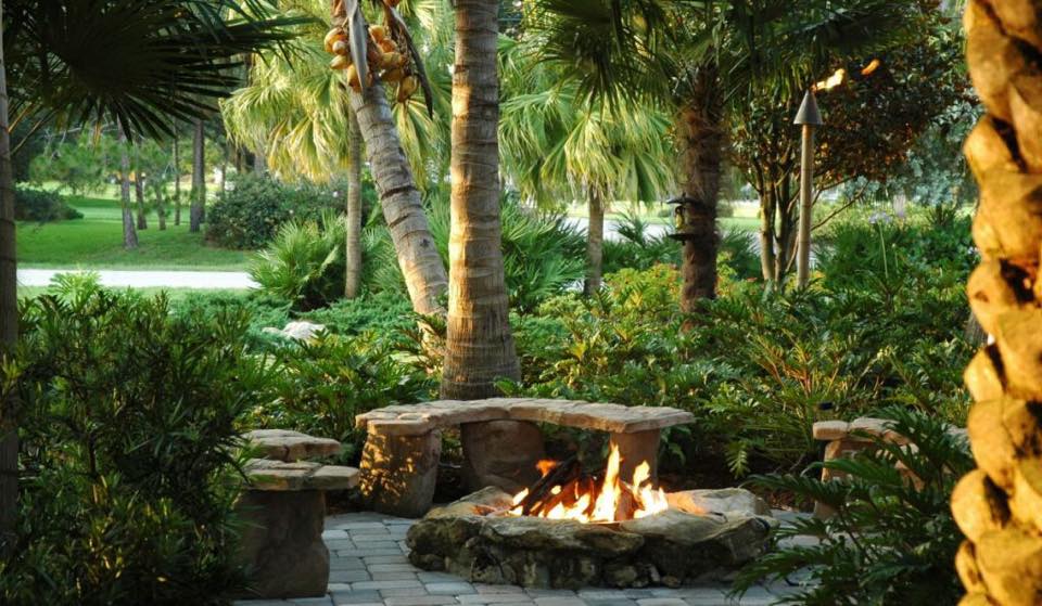 Stone fire pit with benches surrounded by greenery and palm trees.