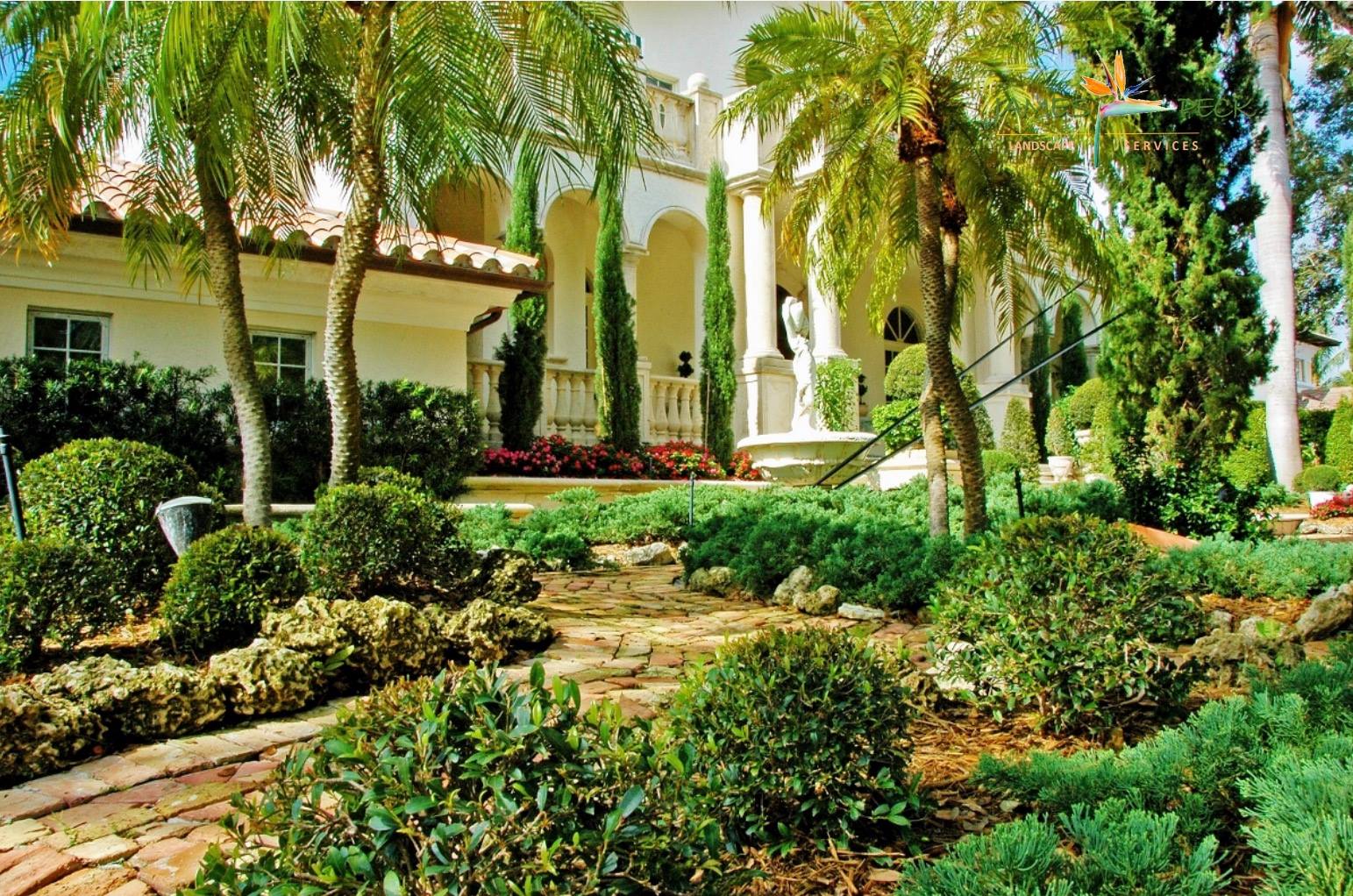 Lush garden with manicured shrubs and palm trees in front of a grand, cream-colored house.