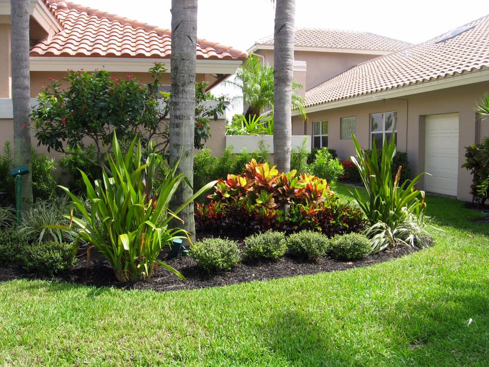 Lush landscaping with green grass, palm trees, and colorful shrubs in front of houses with terracotta tile roofs.