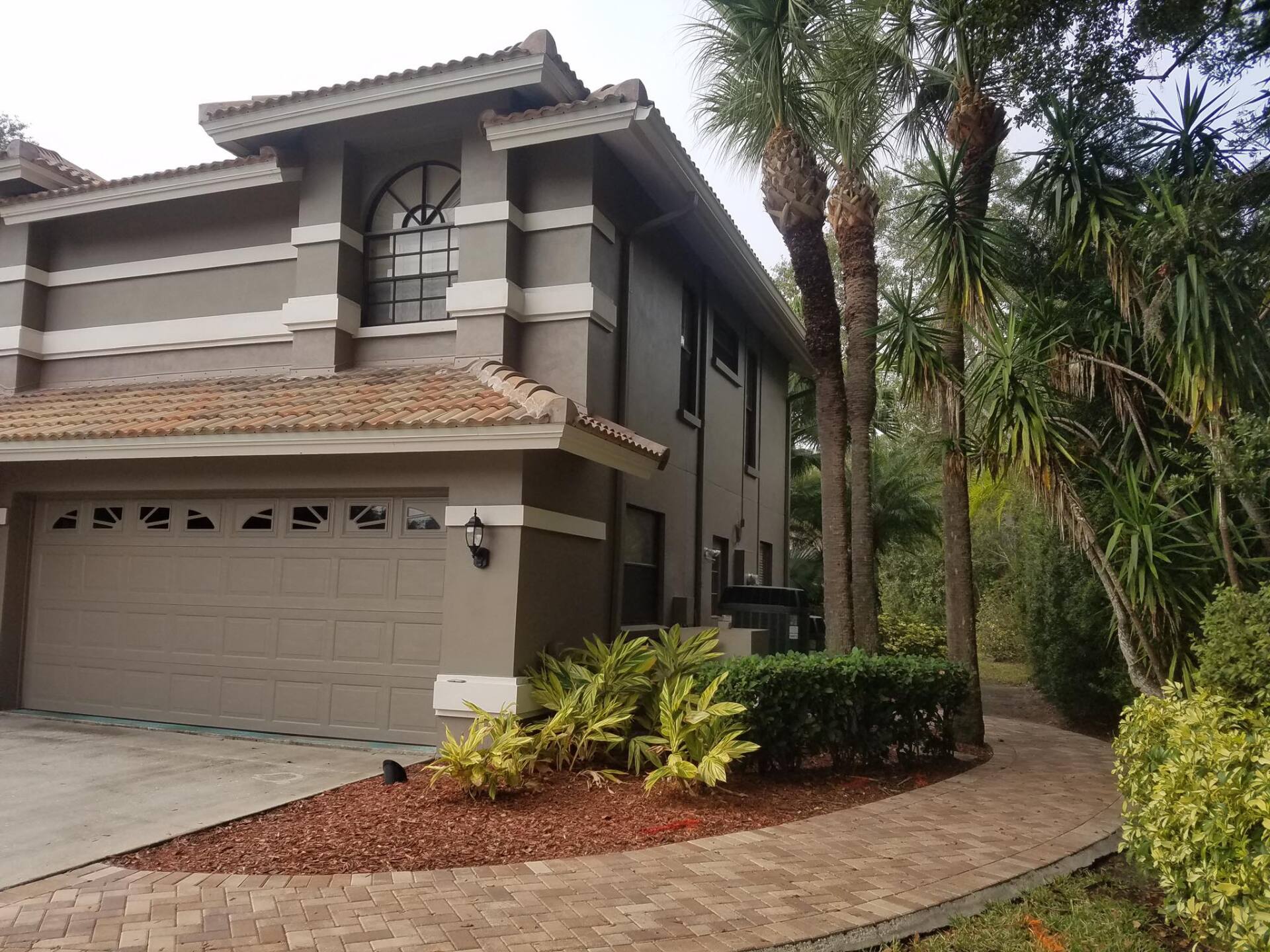 Two-story house with a brown garage door and a brick walkway surrounded by plants.
