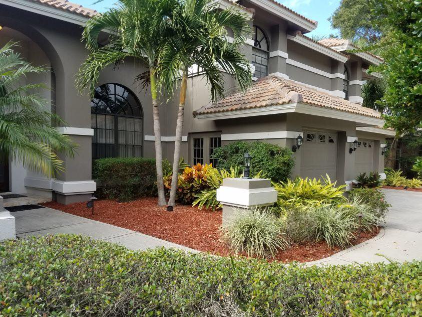 Two-story beige house with tile roof, palm trees, and landscaped front yard with red mulch.