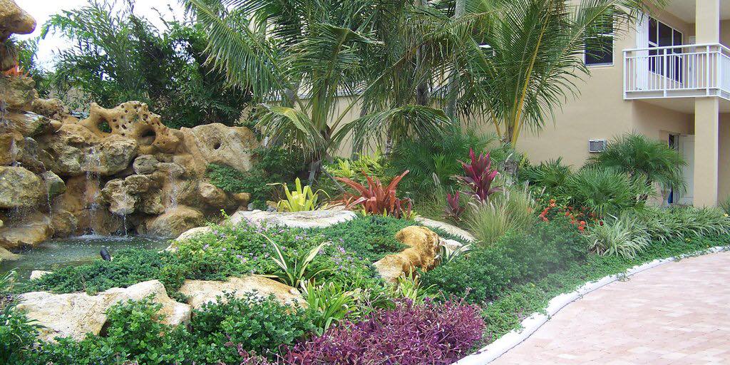 Landscaped garden with waterfall, diverse plants, and a building with a balcony.
