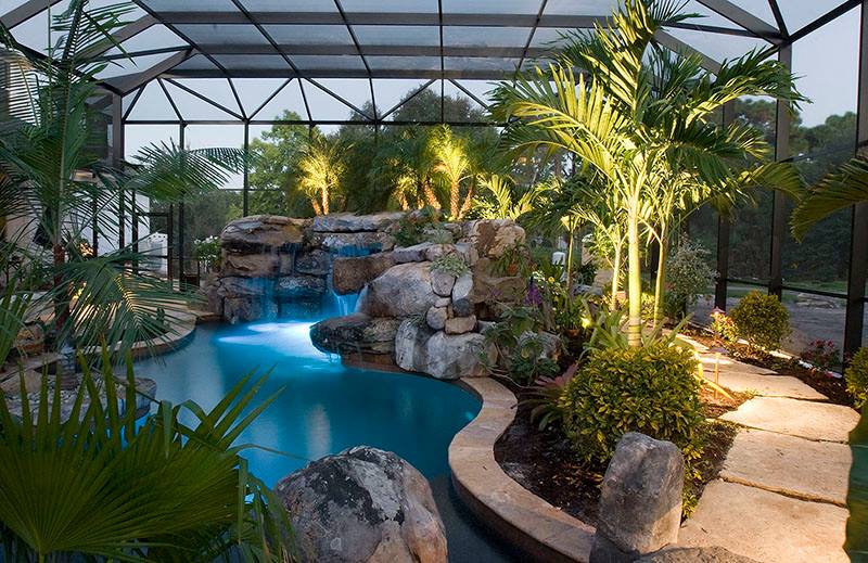 A pool area with a waterfall feature, lush plants, and pathway, all within a screened enclosure.