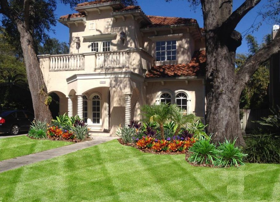 Beige stucco house with a red-tiled roof and lush landscaping; two large trees flank the home.
