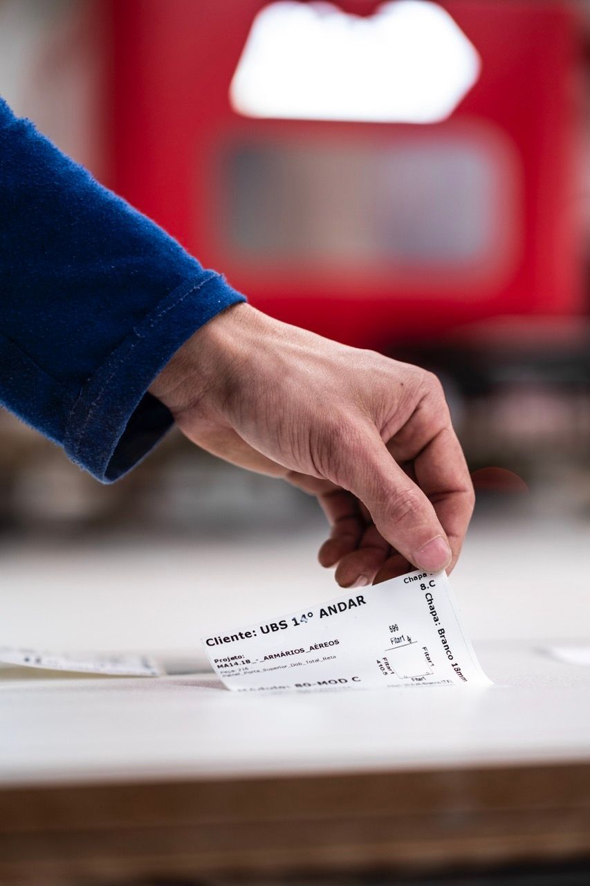 Mão depositando um voto em uma urna transparente com fundo vermelho.