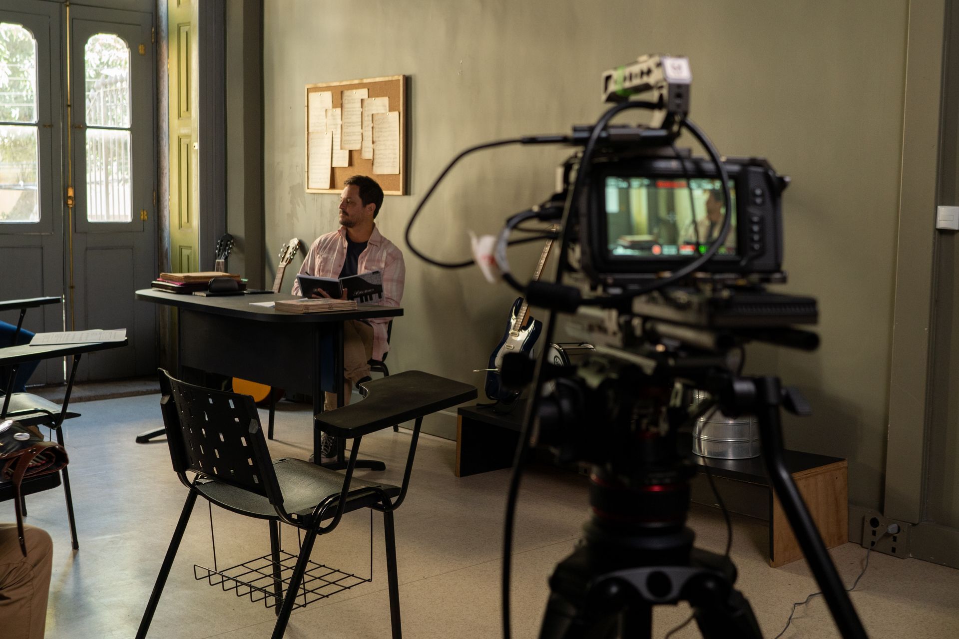 Gravação do curta Let's Jazz A man is sitting at a desk in front of a camera in a classroom.