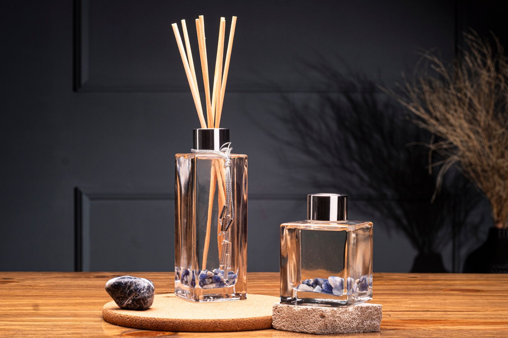 Dois difusores de fragrância em vidro transparente com varetas e um pequeno frasco de perfume sobre uma mesa de madeira.
