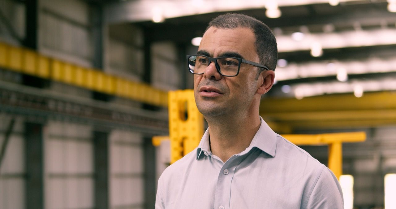 Produção executiva CAF | TAG12 Man wearing glasses in a warehouse, standing near yellow industrial equipment and looking off-camera