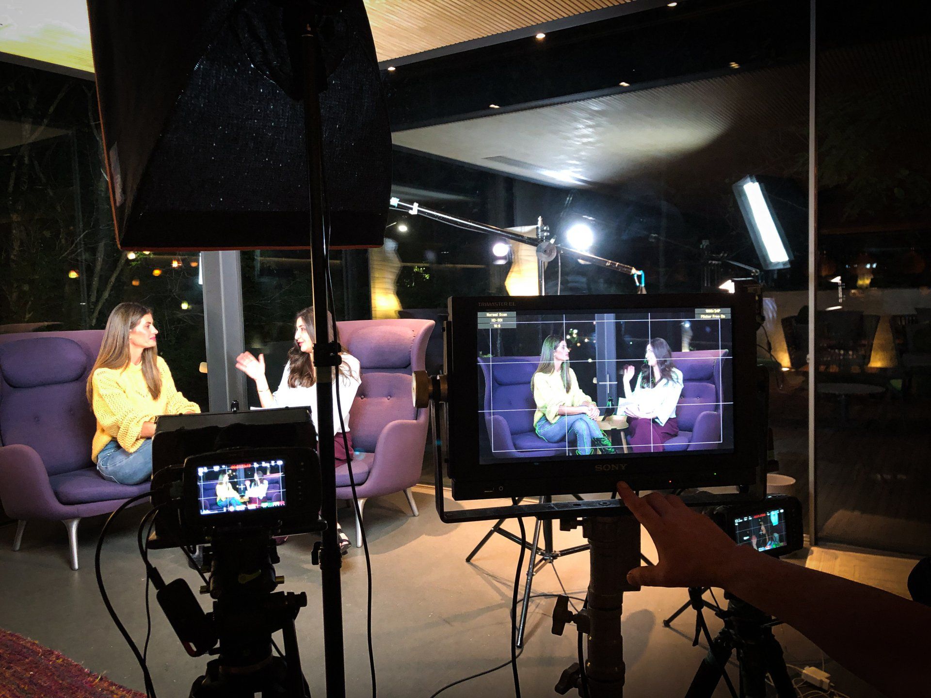 Duas mulheres sendo filmadas em um estúdio com cadeiras roxas. Monitores e equipamentos de iluminação são visíveis.