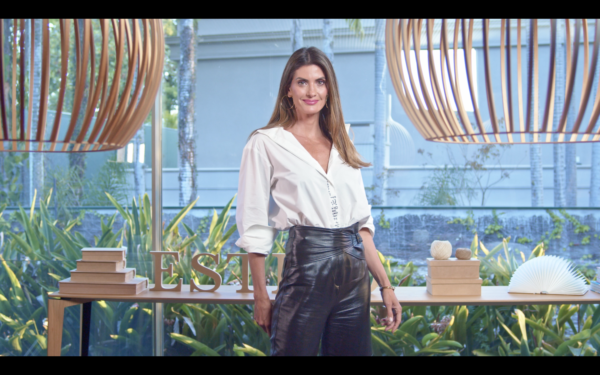 Plano Americano: O Clássico do EAD! Uma mulher de camisa branca e calça de couro preta está parada em frente a uma mesa.