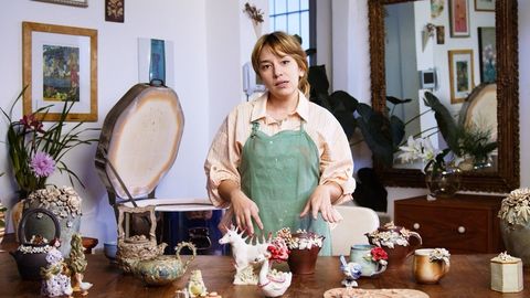 Mulher de avental verde em pé atrás de uma mesa com materiais de artesanato em um quarto aconchegante e decorado.