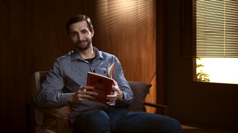 Homem sentado em uma sala com iluminação aconchegante, segurando um livro vermelho aberto junto a uma janela com persianas.