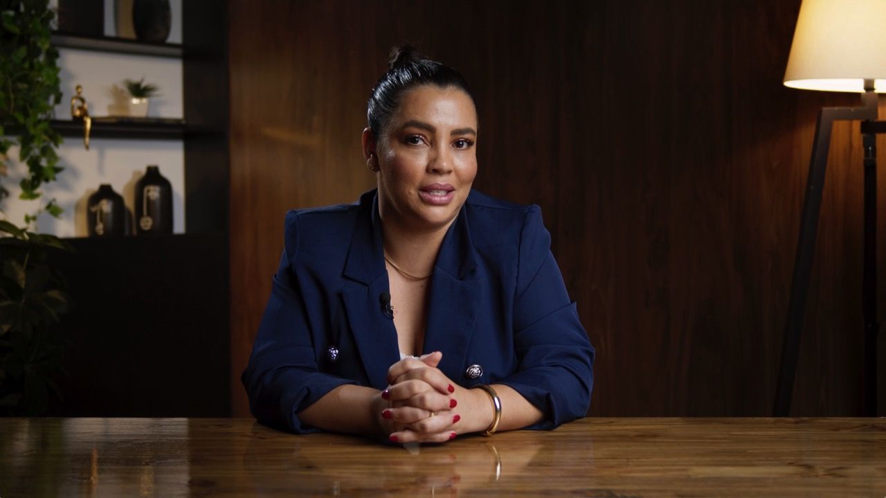 Depoimentos e entrevistas Uma mulher está sentada à mesa com as mãos cruzadas.
